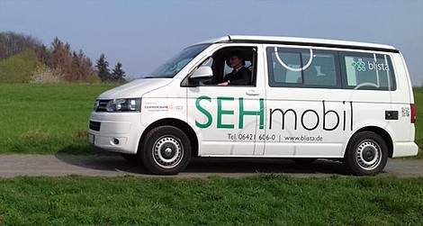 Ein weißer Kleinbus mit der Aufschrift &quot;SEHmobil&quot; fährt auf einer kleinen Straße  von rechts nach links durch das Bild
